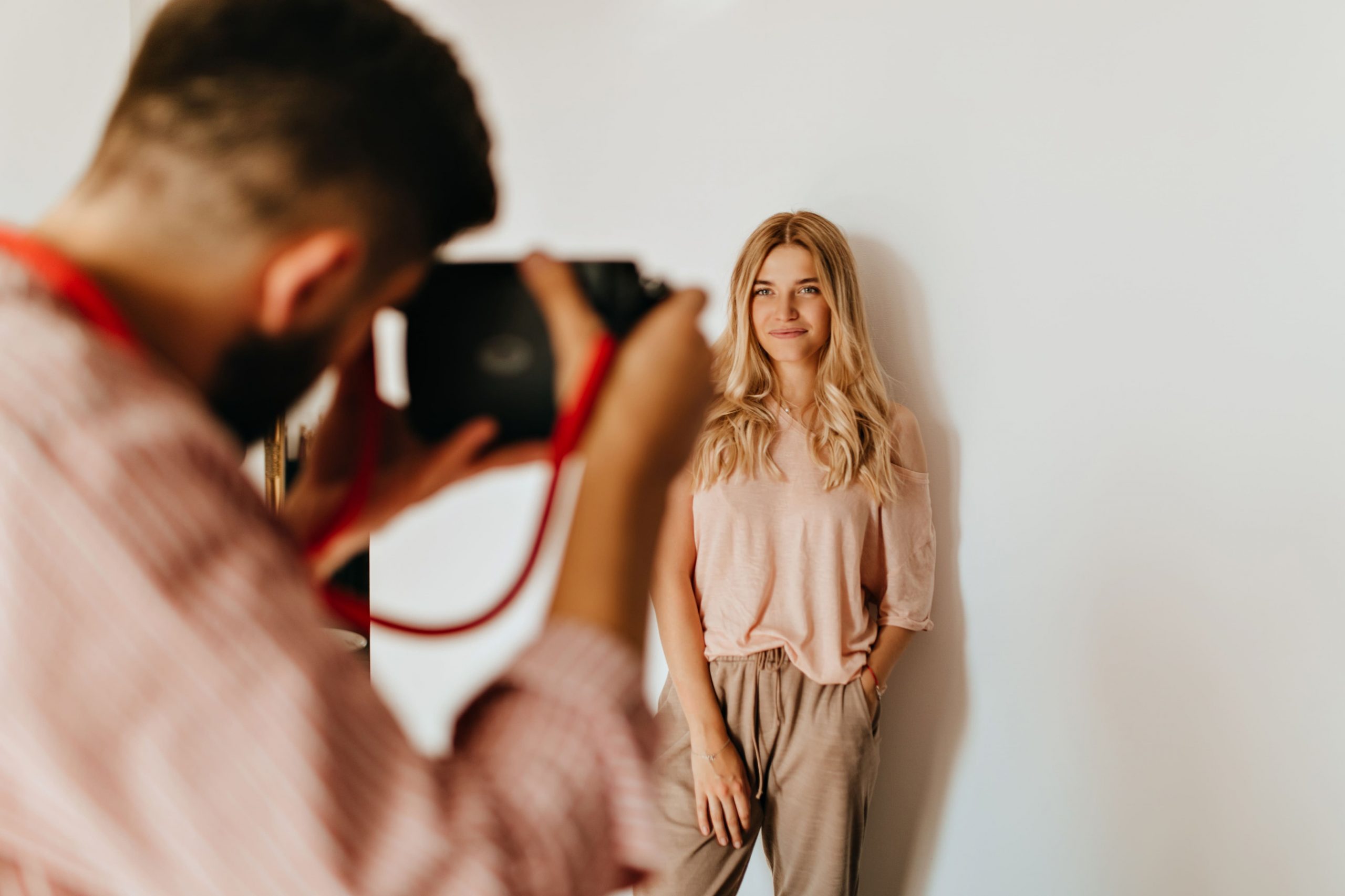 darkhaired-guy-makes-photo-his-blond-girlfriend-dressed-pink-tshirt-beige-pants-against-white-wall
